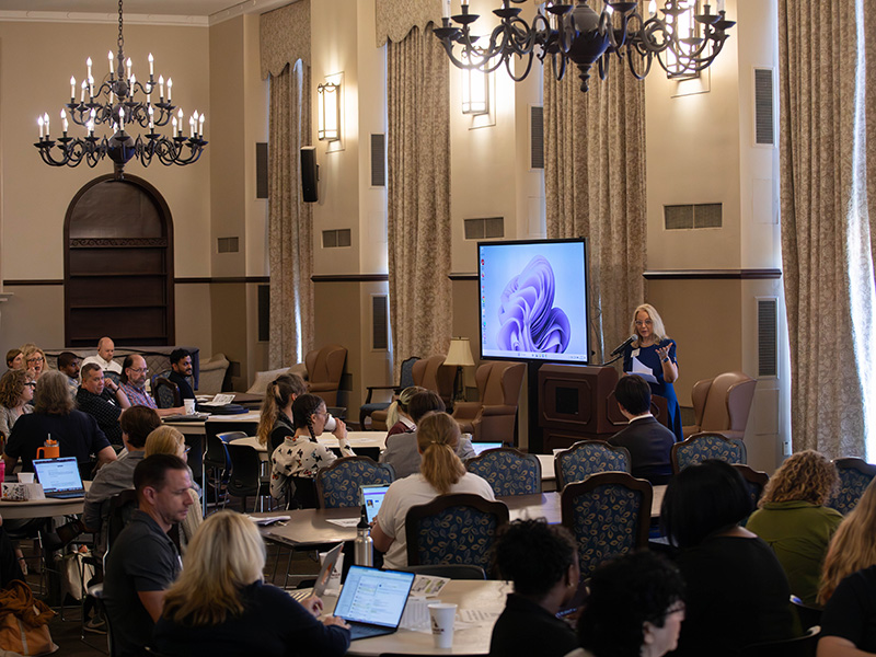A presenter stands at a podium addressing an audience seated at round tables in a chandelier-lit conference room, with a presentation screen displayed at the front.