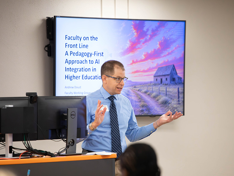 A presenter gestures while speaking beside a computer setup, with a screen behind displaying a presentation titled “Faculty on the Front Line: A Pedagogy-First Approach to AI Integration in Higher Education.”