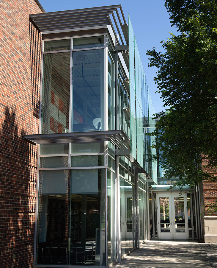 A modern glass and steel entrance structure extends from a brick building, with large windows, metal overhangs and surrounding trees along a sidewalk.