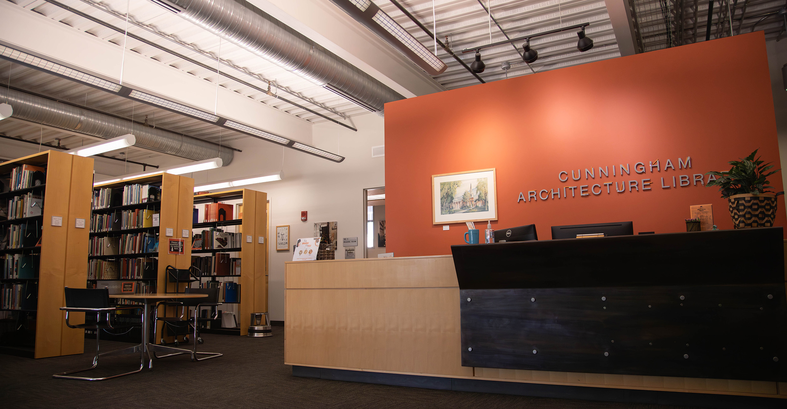 The Cunningham Architecture Library interior features a reception desk, bookshelves filled with volumes, and study tables beneath exposed ceilings and industrial lighting.