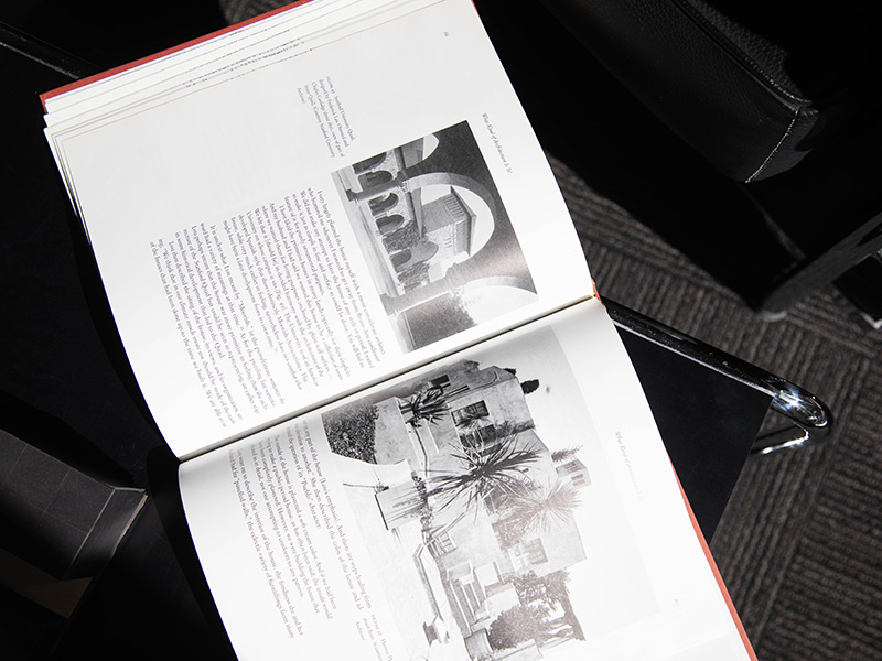 An open book displaying black-and-white architectural photographs and text rests on a table beside modern chairs, highlighting study materials in a library or reading area.
