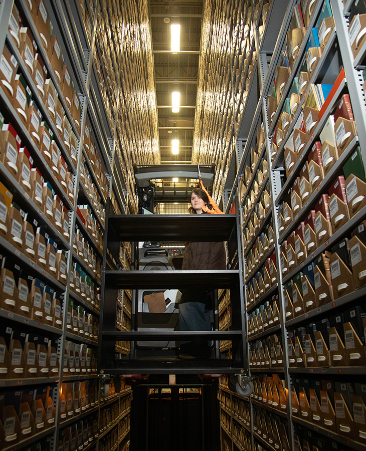 A staff member operates a lift platform between tall rows of archival shelving filled with labeled boxes and books, illustrating the retrieval and management of materials in a high-density library storage facility.