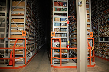 Tall archival library shelving filled with books and labeled storage boxes extends into the distance, illustrating a high-density collection storage area used to preserve and organize library materials.