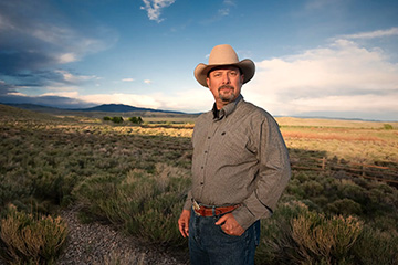 A person wearing a cowboy hat stands in a sagebrush-covered rangeland landscape with distant hills and dramatic clouds in the background.