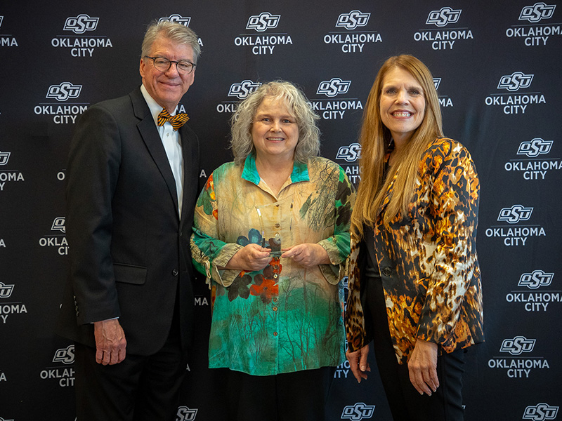 Three adults stand together holding a small glass award and posing for a photo in front of an OSU Oklahoma City–branded backdrop during an employee recognition event.