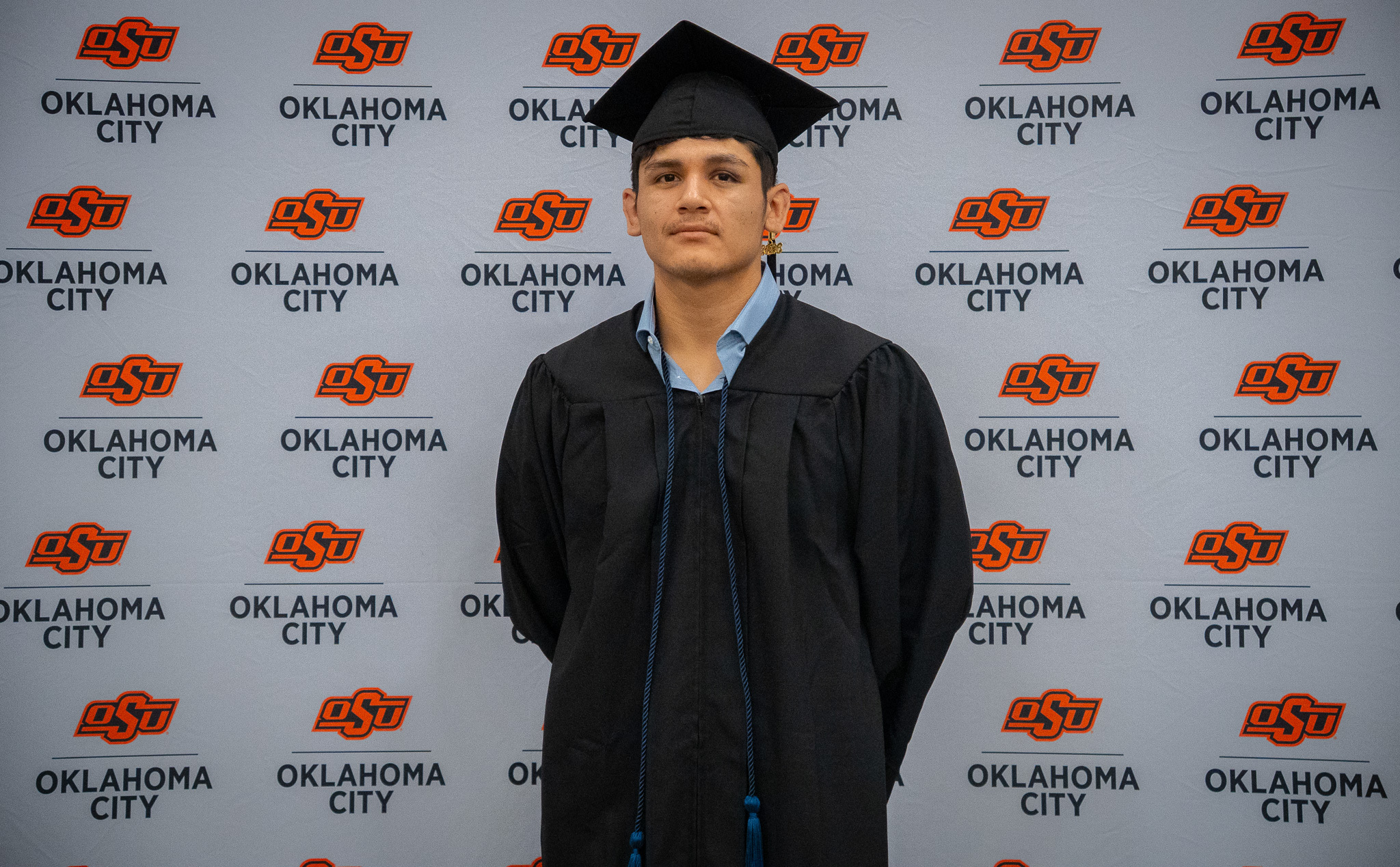 An OSU-OKC graduate wearing a black cap and gown poses in front of an Oklahoma State University-Oklahoma City backdrop during spring commencement ceremonies.