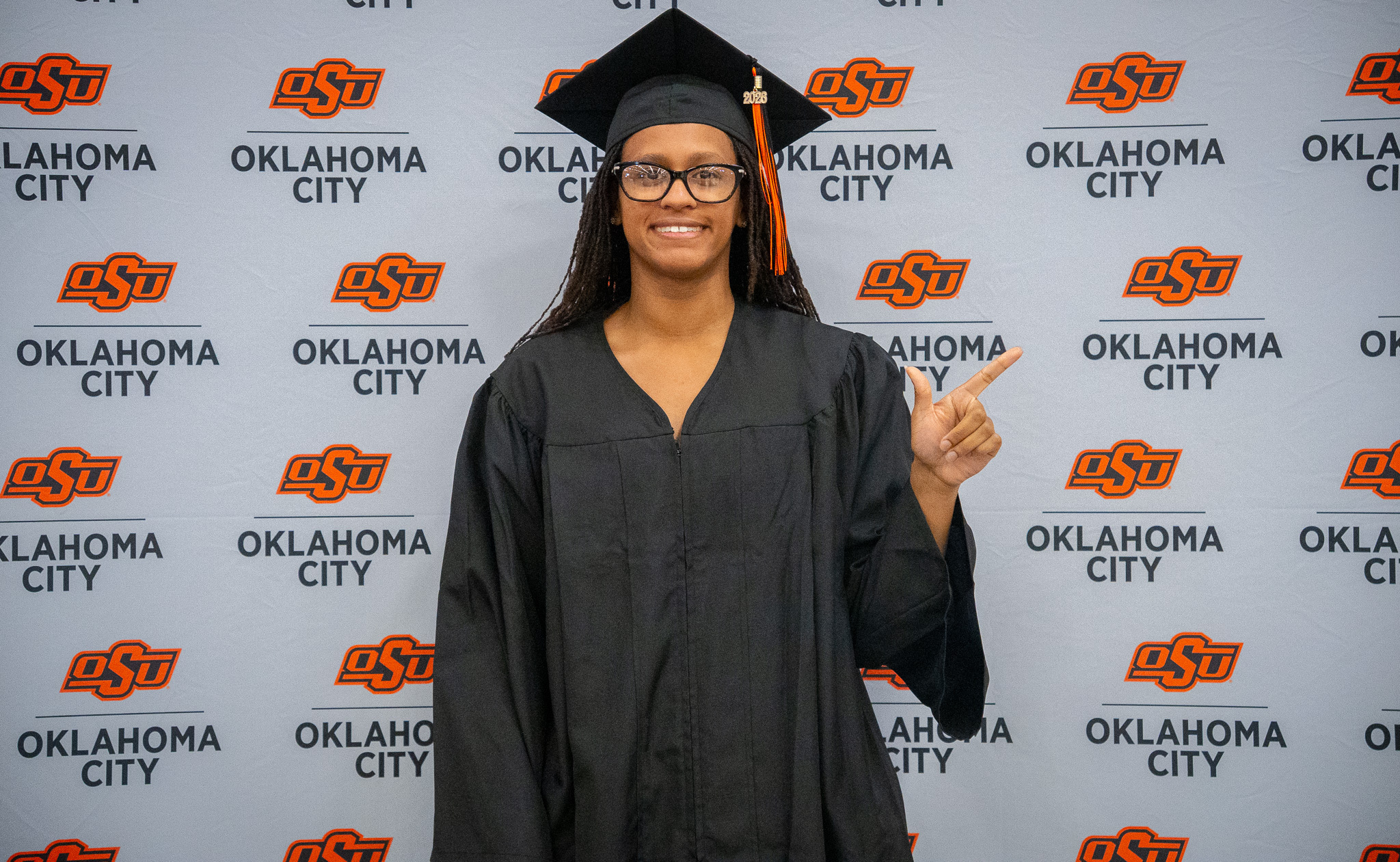 An OSU-OKC graduate wearing a black cap and gown smiles and points while posing in front of an Oklahoma State University-Oklahoma City backdrop during spring commencement ceremonies.