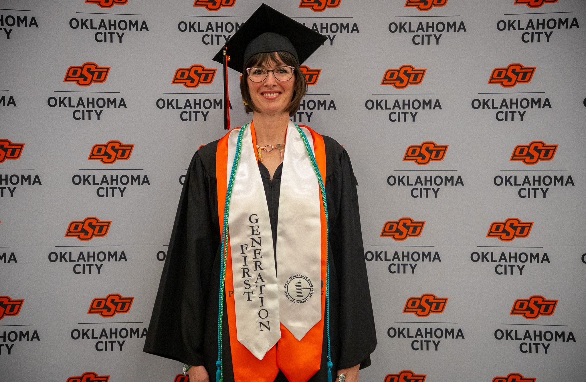 An OSU-OKC graduate wearing a black cap and gown, honor cords and a first-generation stole poses in front of an Oklahoma State University-Oklahoma City backdrop during spring commencement ceremonies.