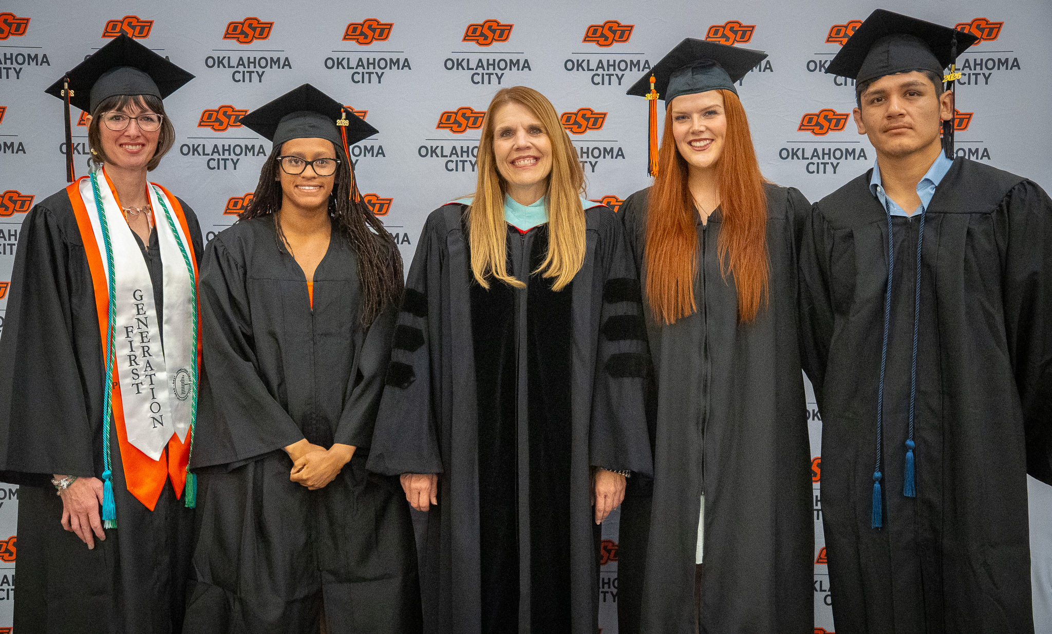 OSU-OKC Outstanding Graduates pose with a faculty member in front of an Oklahoma State University-Oklahoma City backdrop during spring commencement ceremonies. The graduates wear caps and gowns, with one graduate wearing a first-generation stole and honor cords.