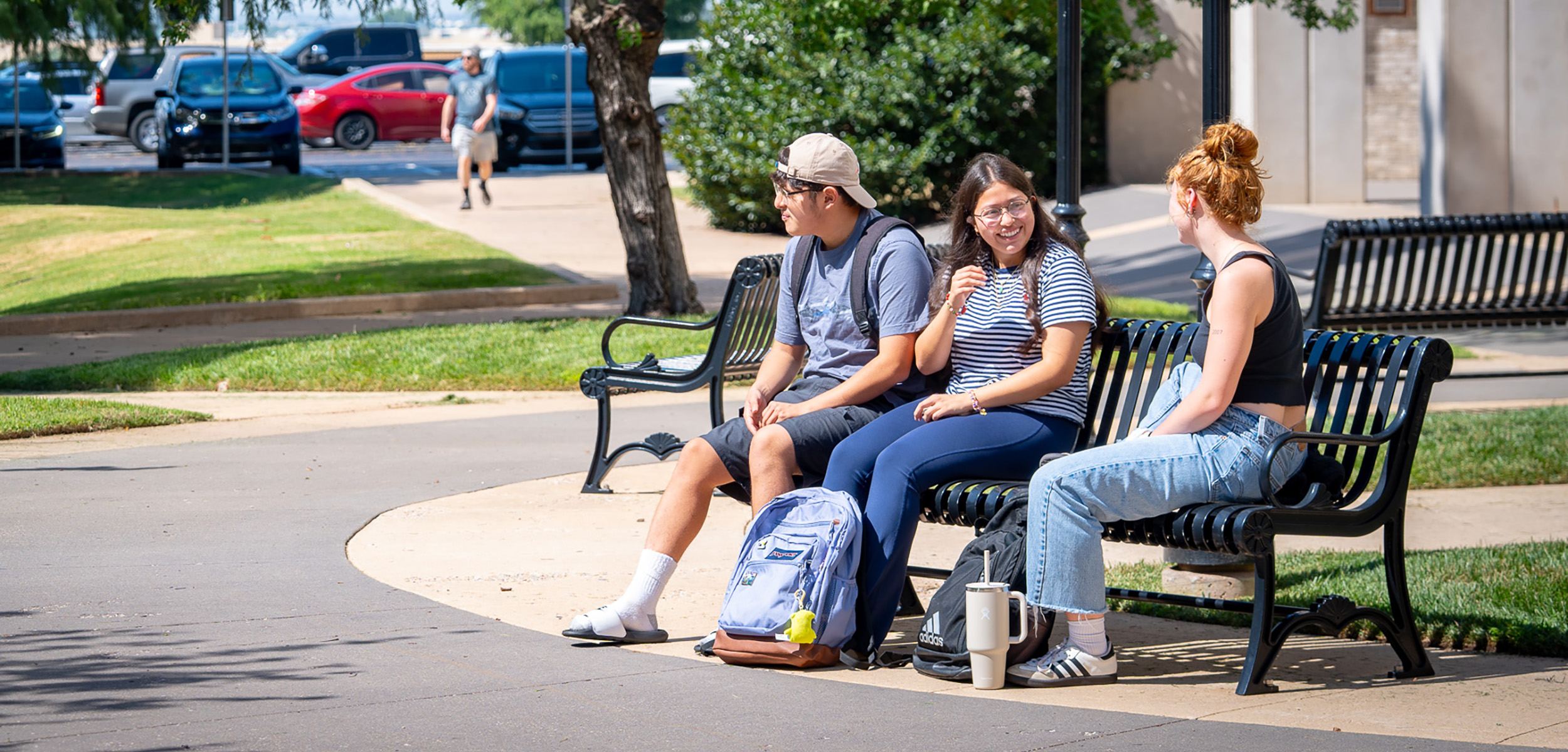 Three students sit on a black bench in a sunny campus courtyard. They are chatting and smiling, with backpacks and tumblers beside them, conveying a relaxed and cheerful atmosphere.