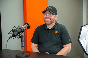 A man wearing an Oklahoma State University polo shirt and cap sits at a table in a recording studio, smiling during a podcast or interview session. A microphone is positioned in front of him, and studio lighting equipment is visible nearby.