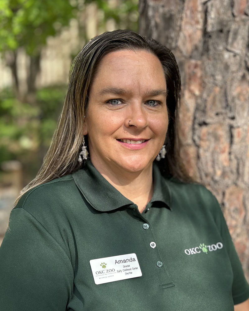 Portrait of a person wearing a green OKC Zoo polo shirt and name badge, standing outdoors near a tree in a natural setting.