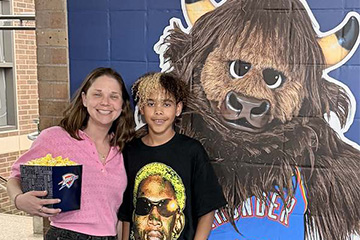 An adult and a child stand smiling together in front of a colorful bison mural. The adult holds a blue notebook, and the child wears a graphic T-shirt. The photo appears to have been taken inside a school or community building.