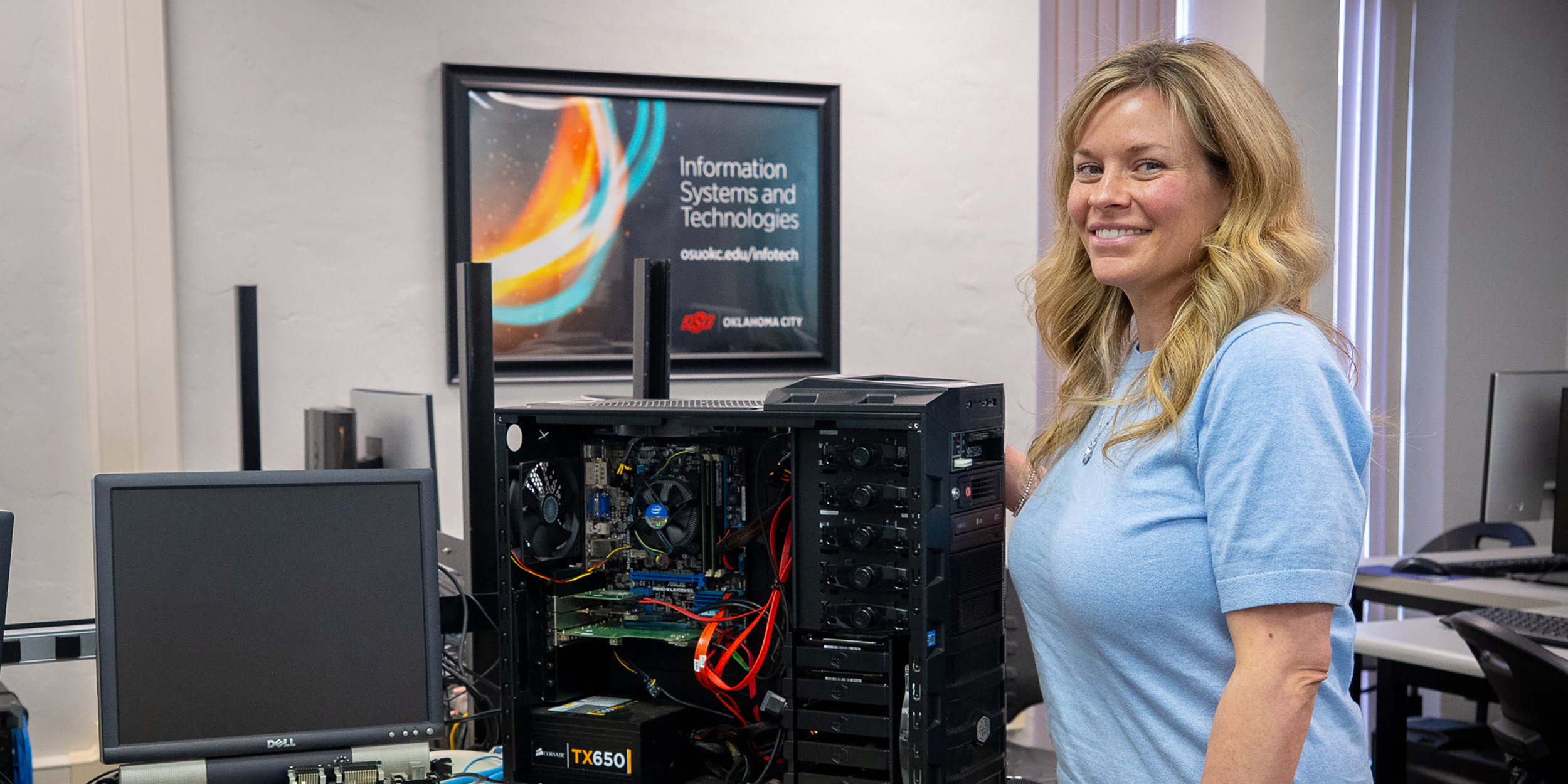 A woman in a light blue shirt stands smiling next to an open computer tower in a tech room. A poster reads "Information Systems and Technologies."