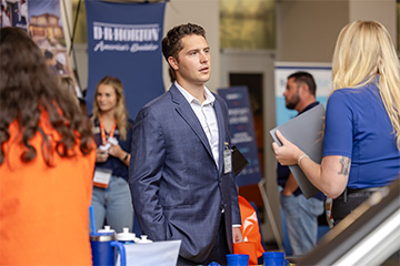 A student in a suit speaks with recruiters during a career fair event as attendees network in the background.