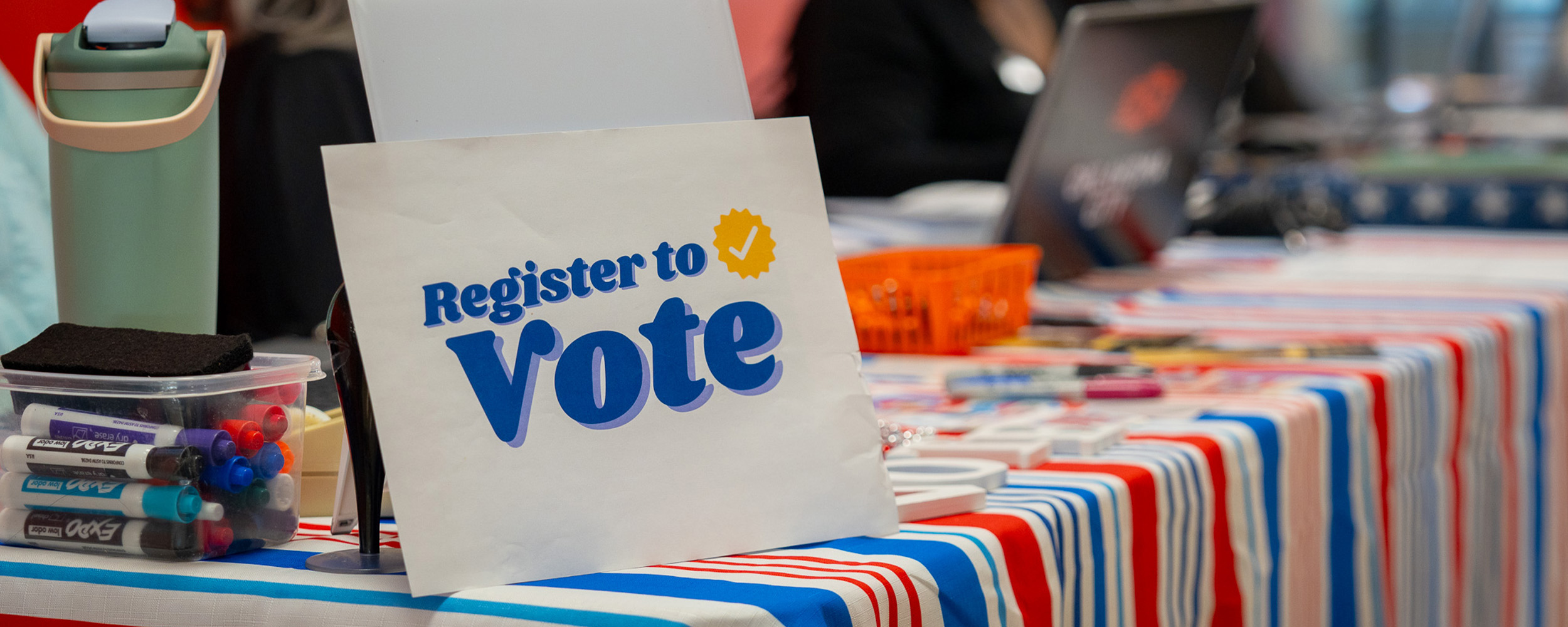A voter registration table with a “Register to Vote” sign, markers and election materials on a red, white and blue tablecloth.