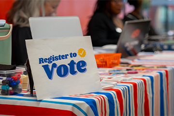 A voter registration table with a “Register to Vote” sign, markers and election materials on a red, white and blue tablecloth.