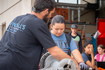 An instructor demonstrates the use of industrial equipment to a student wearing work gloves during a hands-on training activity. Several students watch and observe in a workshop setting with tools and machinery visible in the background.