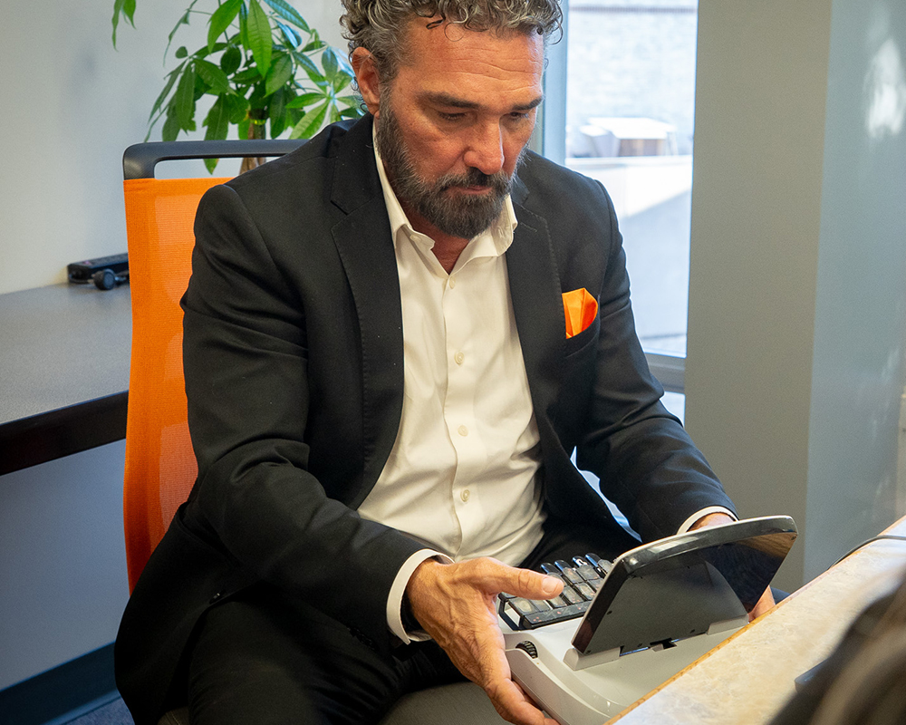 A man in a suit sits in an office on an orange chair, focused on using a stenotype machine. The scene conveys concentration.