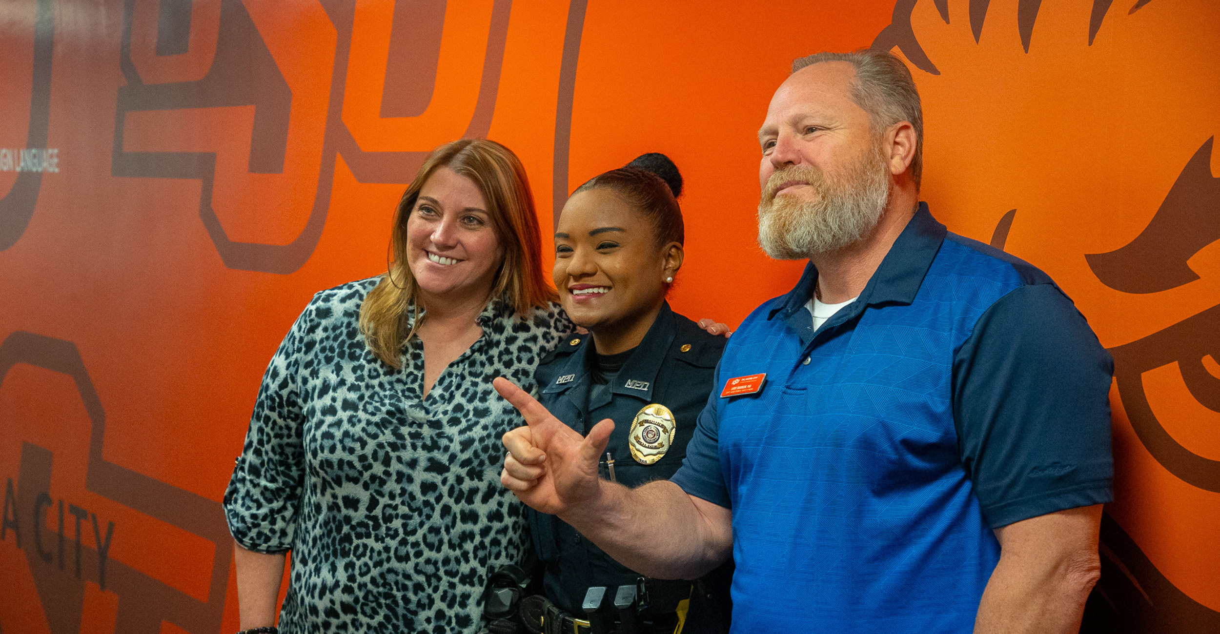 Three people stand close together for a posed photo in front of an orange background, with one person wearing a dark uniform and badge centered between two casually dressed individuals.