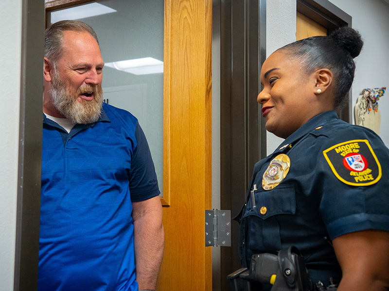 A person wearing a police uniform stands in a doorway speaking with another person in a blue shirt inside an office hallway, with a wooden door frame, visible badge and shoulder patch, and office décor in the background.