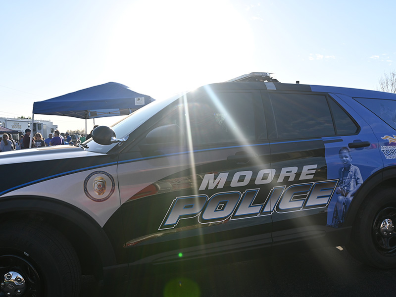 A Moore Police SUV is parked outdoors at a community event, with the large “MOORE POLICE” lettering and department graphics clearly visible on the side as people and tents appear in the background.