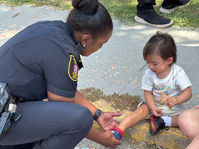 A uniformed police officer kneels on a sidewalk to assist a small child seated on the ground, adjusting the child’s shoe or ankle while debris and grass are visible around them, illustrating a moment of community care.