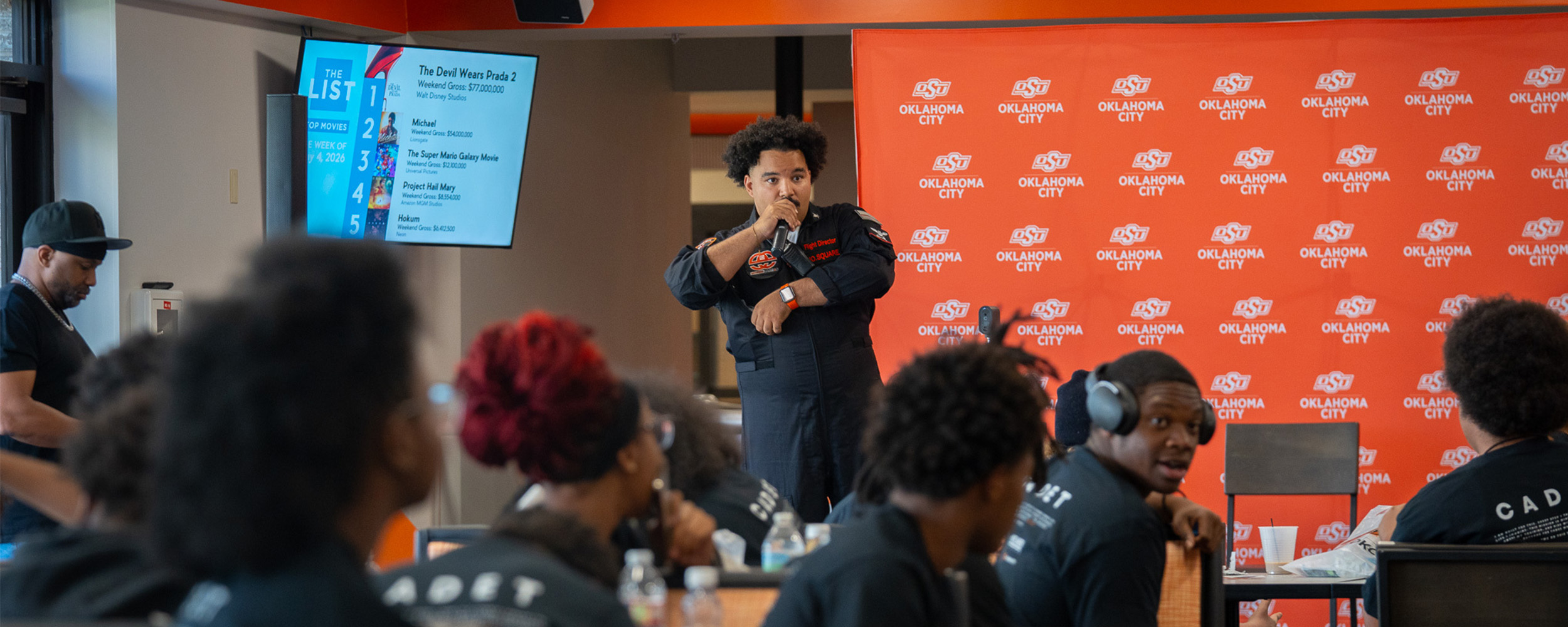 An Oklahoma State University-Oklahoma City speaker addresses students during a campus event as attendees listen from tables in front of an OSU-OKC backdrop.