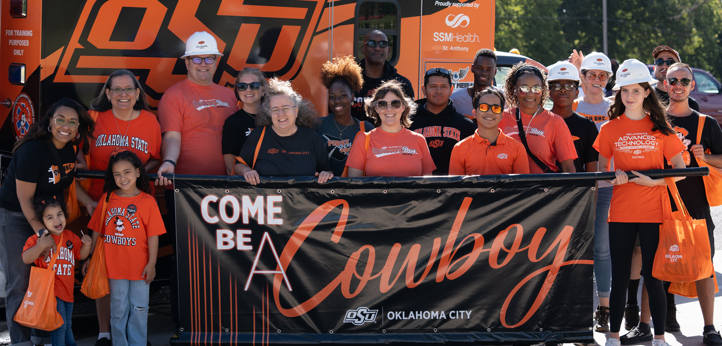Group of Oklahoma State University students, staff, and families wearing orange and black OSU shirts and hard hats, standing together holding a banner that reads "Come Be A Cowboy – OSU Oklahoma City.