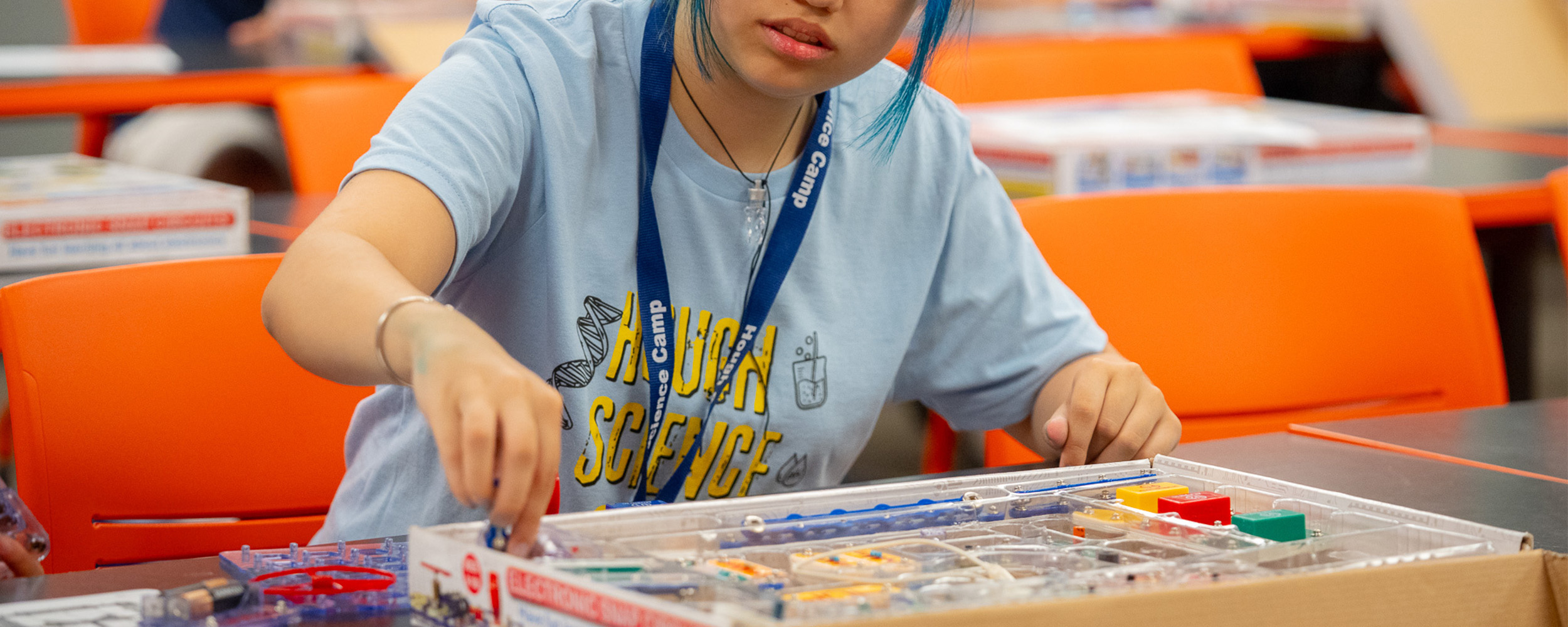 A student participates in a hands-on science activity, assembling components from an educational electronics kit at a classroom table. The student wears a camp T-shirt and lanyard, while other participants work on similar projects in the background.