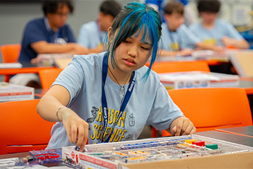 A student participates in a hands-on science activity, assembling components from an educational electronics kit at a classroom table. The student wears a camp T-shirt and lanyard, while other participants work on similar projects in the background.