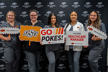 Five Oklahoma State University-Oklahoma City students pose together in front of an OSU-OKC backdrop, holding school-themed signs, including “Go Pokes!” and “Future Healthcare Manager.”
