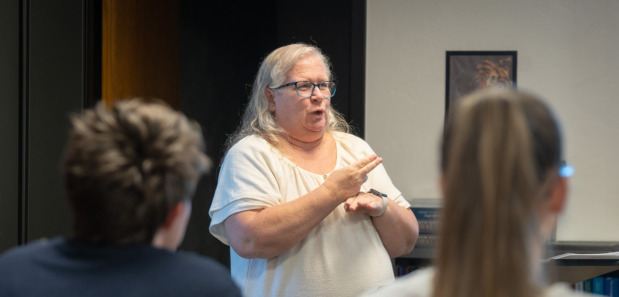 A woman with glasses is signing in a classroom, engaging two students in the foreground. The scene conveys a focused and educational atmosphere.