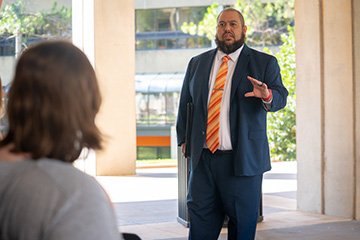 A man in a suit and striped orange tie speaks to a seated audience member in an open indoor space. He gestures with one hand while holding a folder at his side, appearing to lead a presentation or discussion.