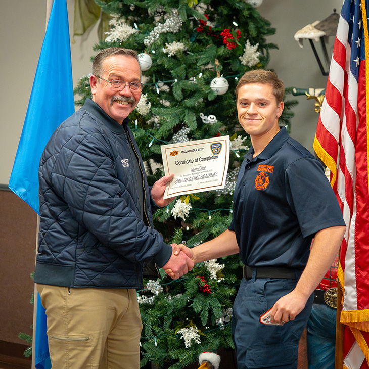 Two uniformed individuals shake hands while holding a certificate of completion in front of a decorated Christmas tree, marking the successful completion of a fire or safety training program.