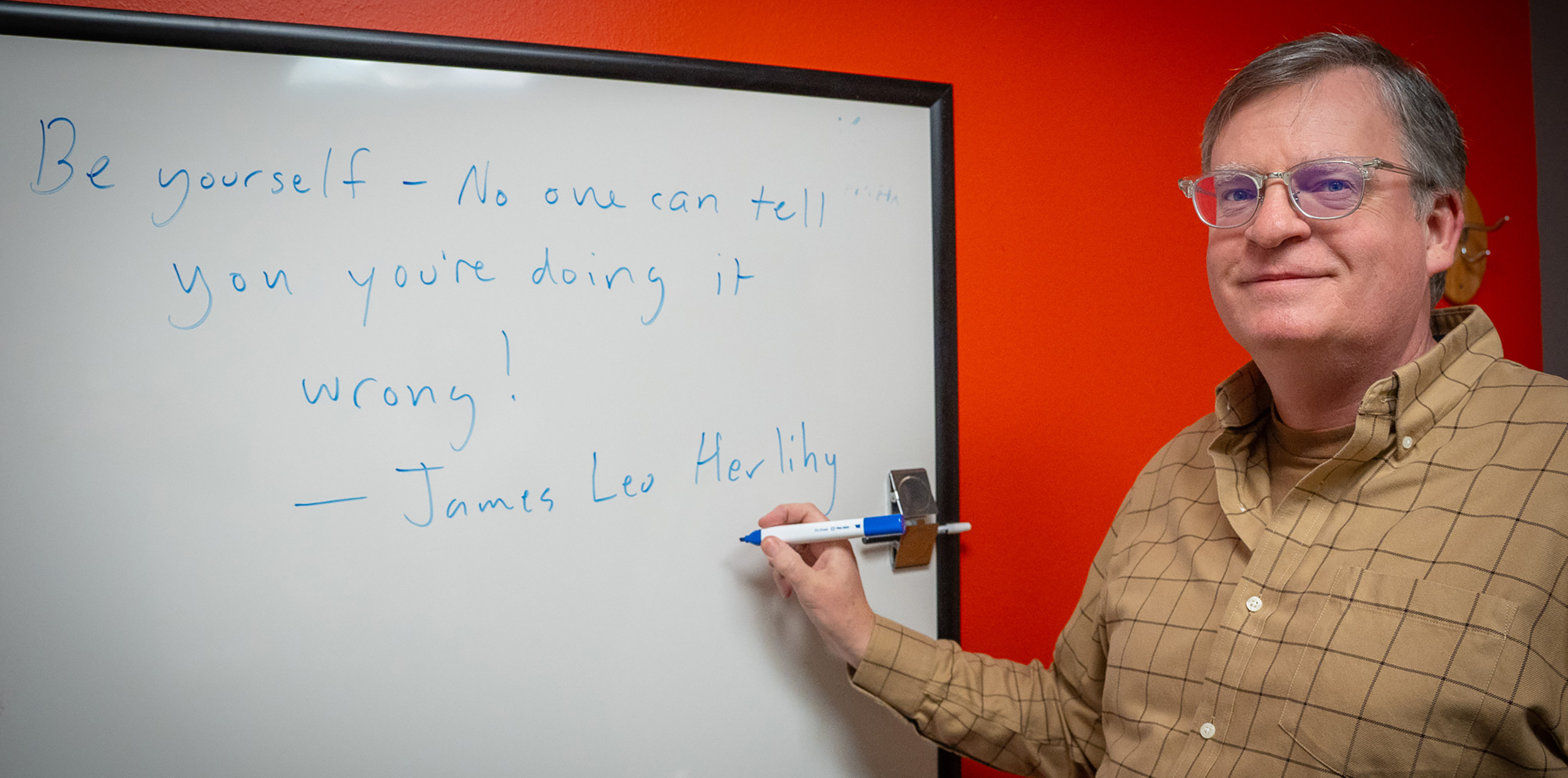 Man in a plaid shirt stands smiling at a whiteboard with a motivational quote about being yourself. The background is bright red, conveying positivity.