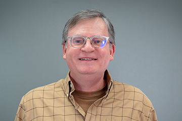 Man in a plaid shirt stands smiling at a whiteboard with a motivational quote about being yourself. The background is bright red, conveying positivity.