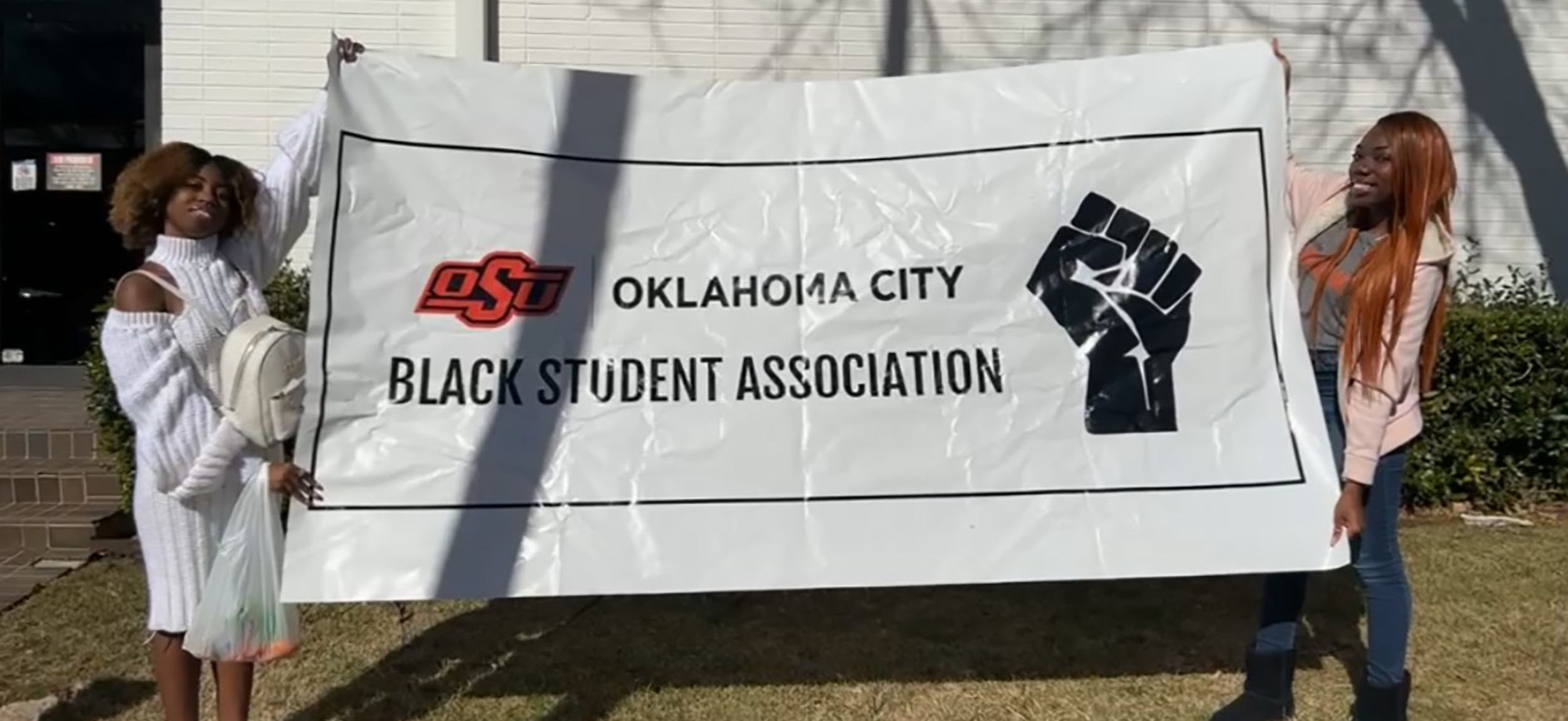 Two people hold a large banner reading "Oklahoma City Black Student Association" with an OSU logo and a black fist. The mood is proud and uplifting.