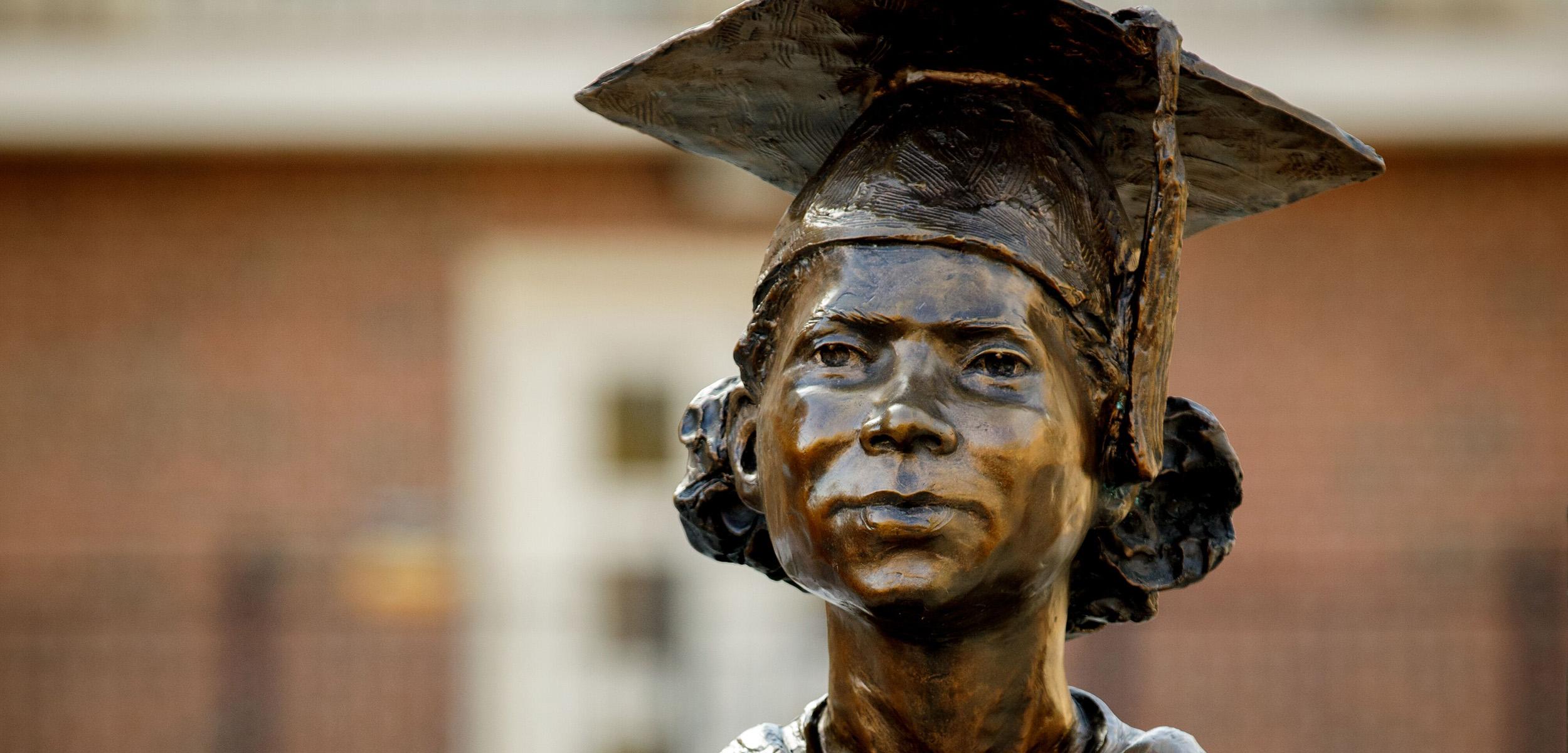 Bronze statue of a young Nancy Randolph Davis wearing a graduation cap. The expression is thoughtful and proud. A blurred brick building is in the background.
