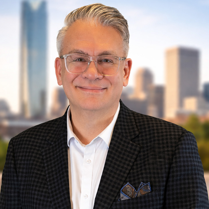 A professional head-and-shoulders portrait of a person wearing glasses, a white shirt, and a dark jacket, photographed against a softly blurred city skyline background.