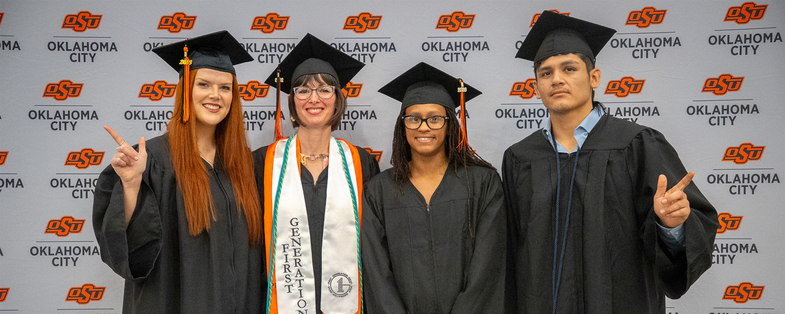 Four OSU-OKC graduates pose together in caps and gowns in front of an Oklahoma State University-Oklahoma City backdrop during spring commencement ceremonies. One graduate wears an “Outstanding Graduate” stole while the group smiles and celebrates the occasion.