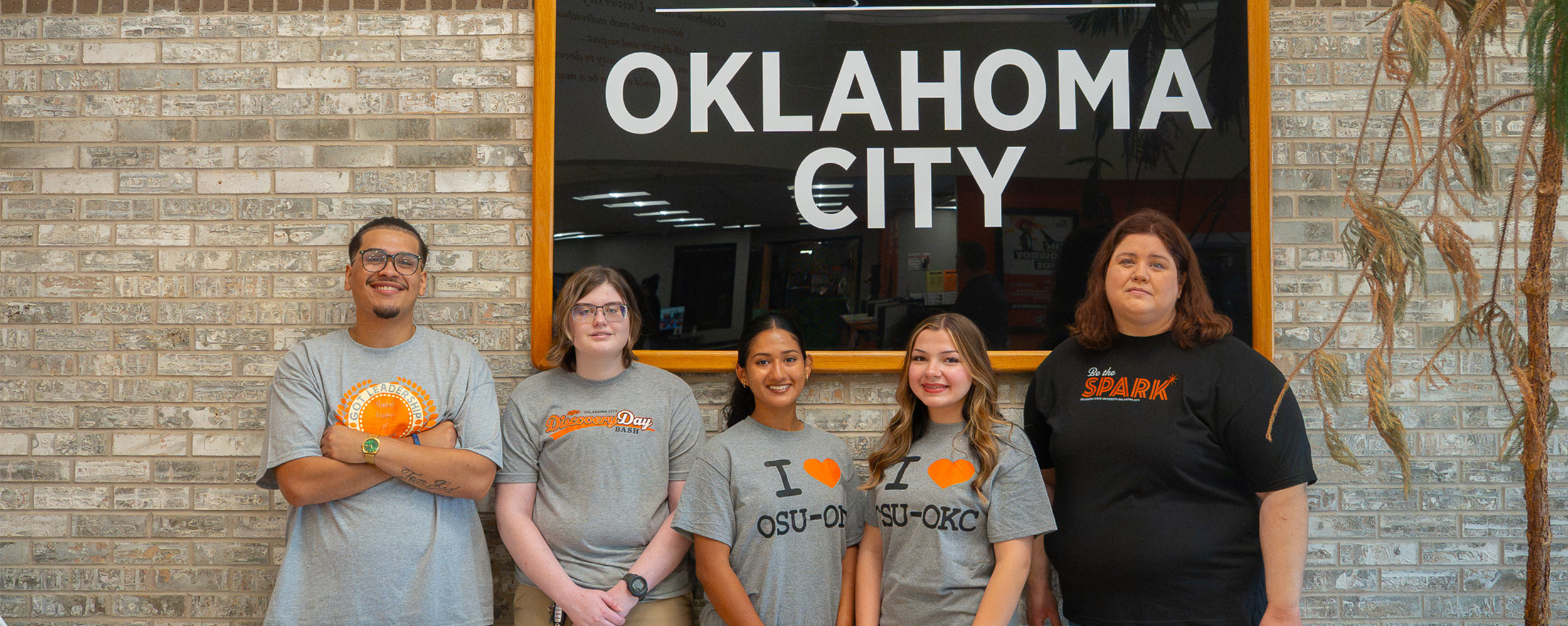 Five students stand together in front of an Oklahoma State University-Oklahoma City sign, wearing OSU-OKC apparel and posing for a group photo.