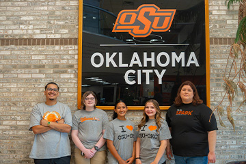 Five students stand together in front of an Oklahoma State University-Oklahoma City sign, wearing OSU-OKC apparel and posing for a group photo.