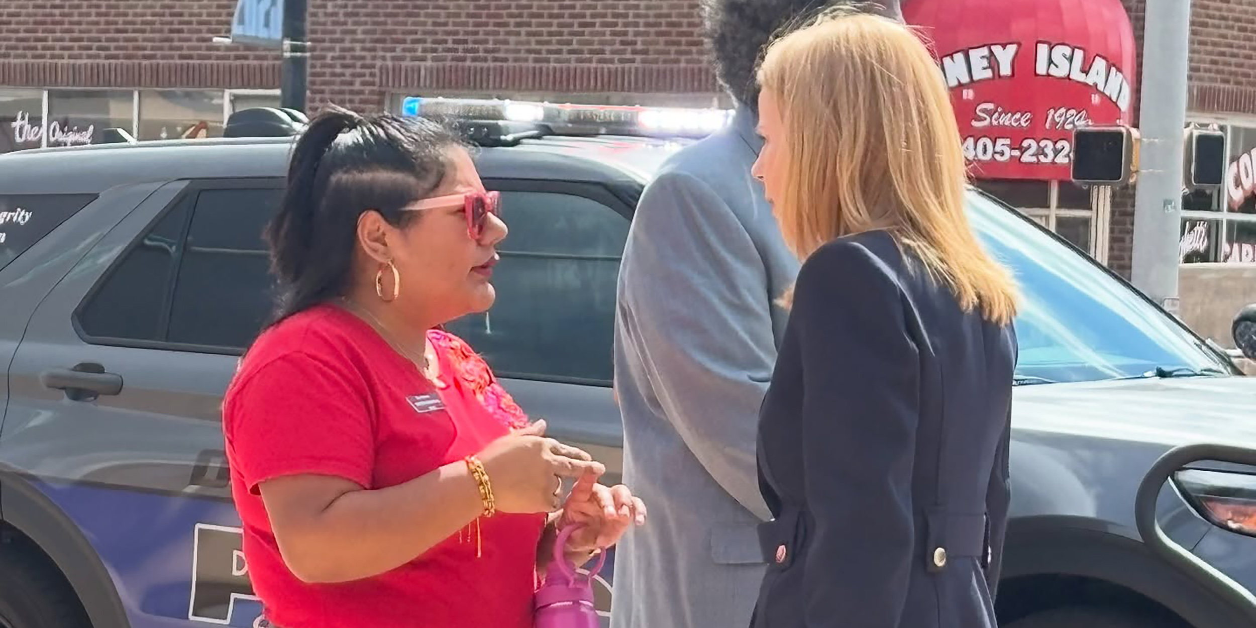 Three people converse on a city sidewalk near a parked police vehicle and storefronts, capturing an informal interaction during a community or campus outreach event.