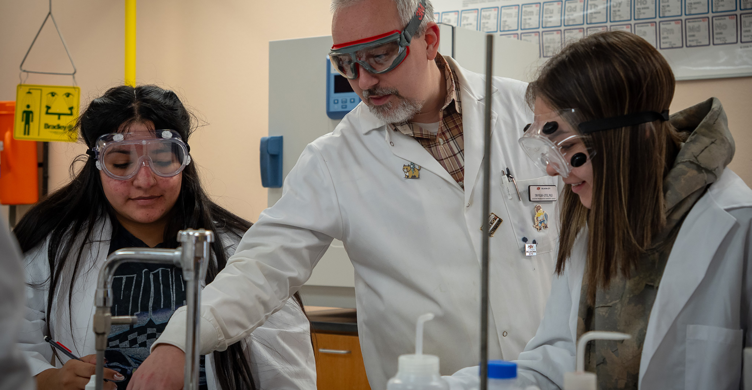 A teacher in a lab coat and goggles guides two students, also in goggles, through a science experiment in a lab, fostering focus and collaboration.