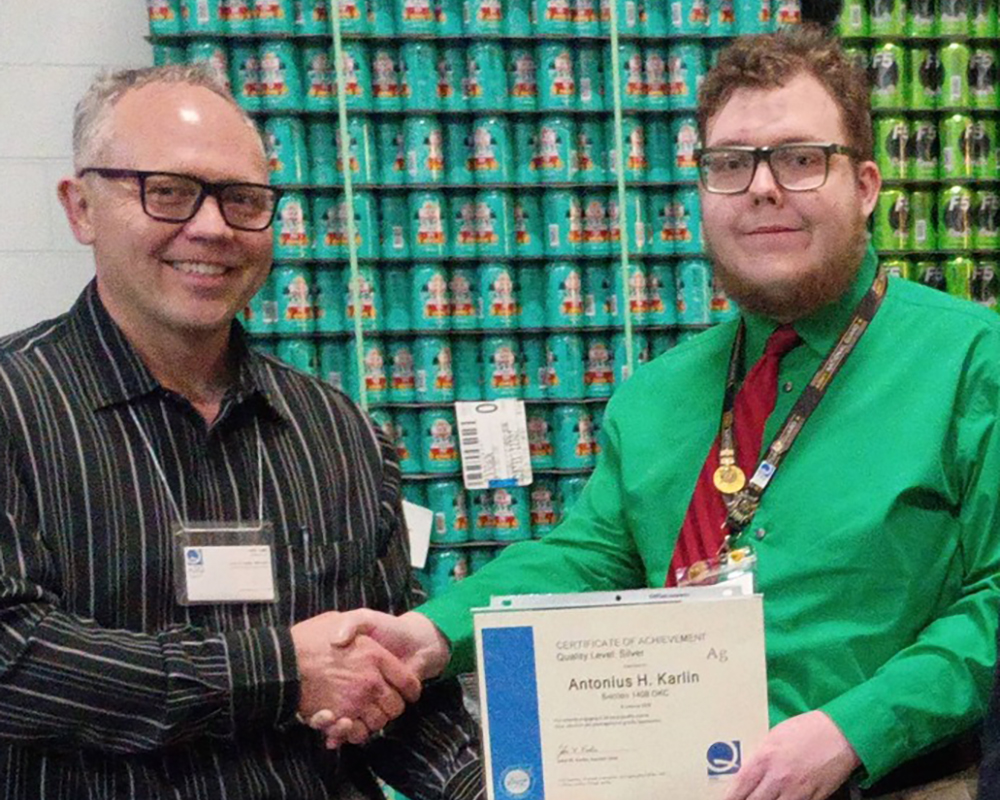 Two men wearing glasses shake hands while holding a certificate of achievement. One wears a striped shirt and name badge, and the other wears a green shirt with a red tie and lanyard. Behind them is a wall stacked with rows of canned beverages.