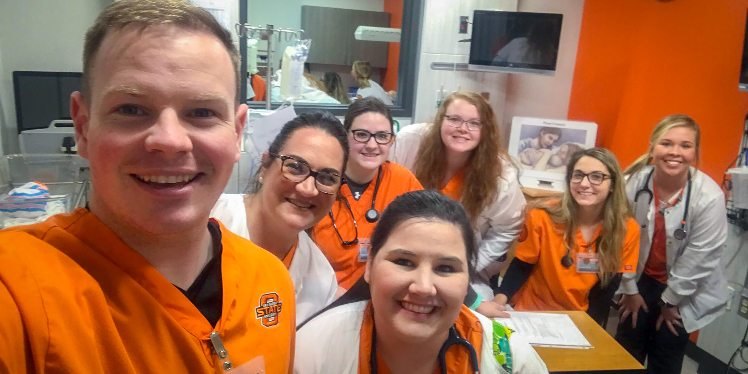 A group of seven smiling nurses, wearing orange and white uniforms, gather for a cheerful selfie in a medical setting with orange walls.