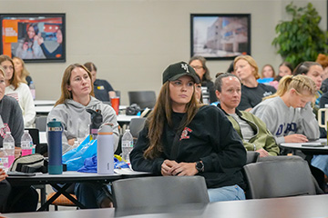 A group of attendees sits at tables in a classroom-style setting, listening to a presentation. Participants take notes and look toward an off-camera speaker, with water bottles, notebooks and personal items on the tables. Framed photos hang on the