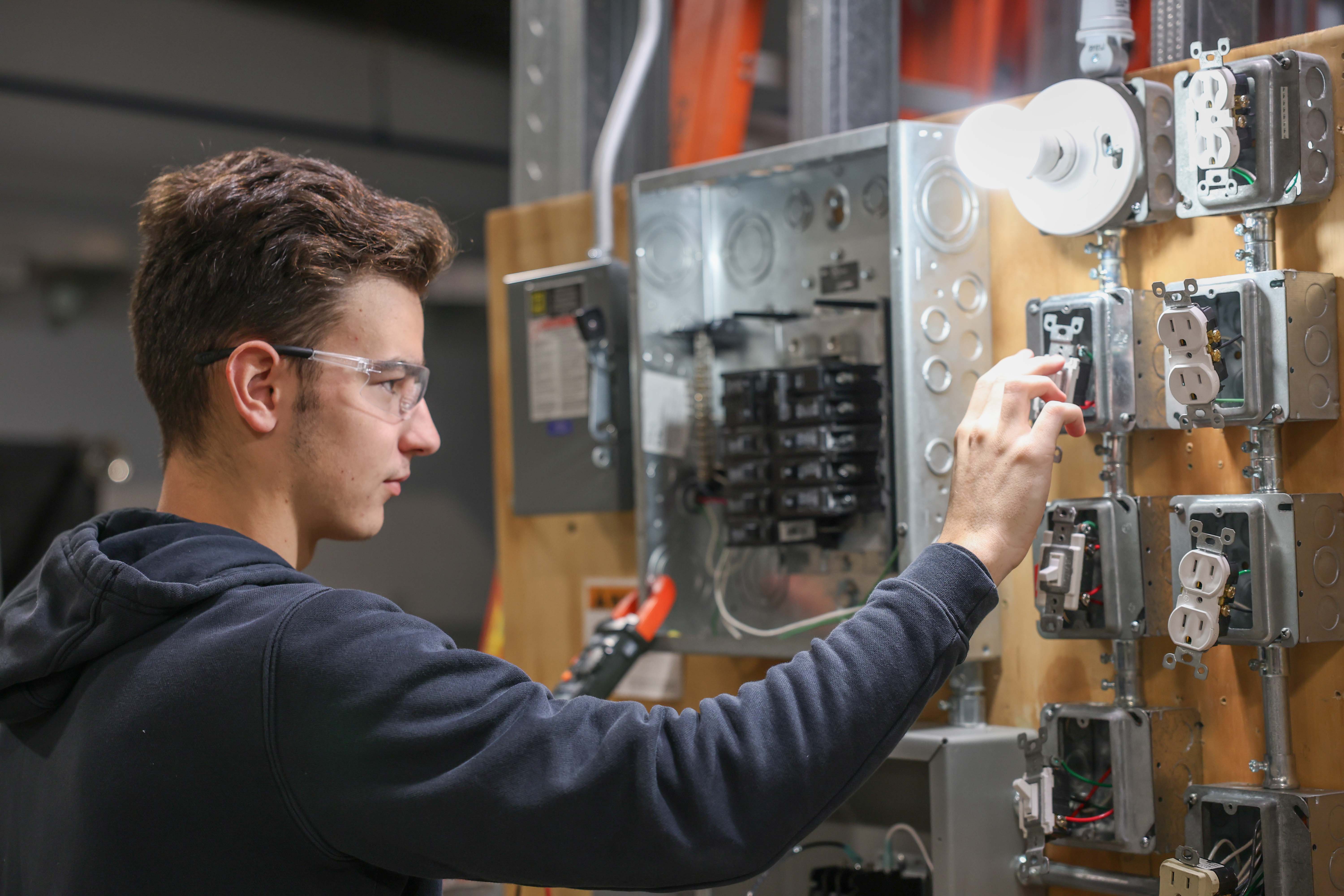 A student wearing safety glasses works at an electrical training wall, wiring a light switch and outlets on mounted metal electrical boxes during a hands-on lab.
