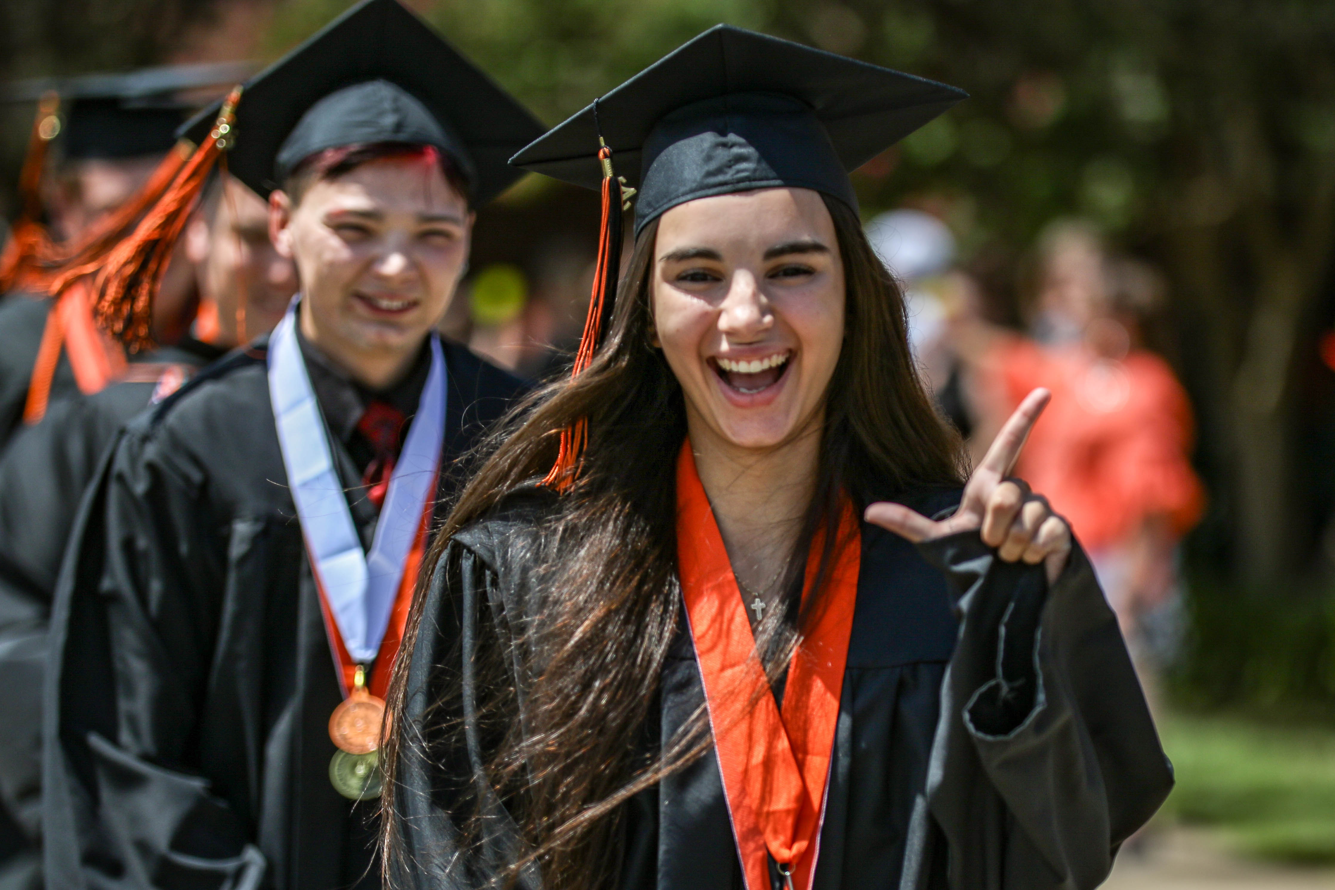 A smiling OSUIT graduate wearing a black cap and gown with an orange stole gestures excitedly toward the camera while walking in a commencement procession, with other graduates following behind outdoors.